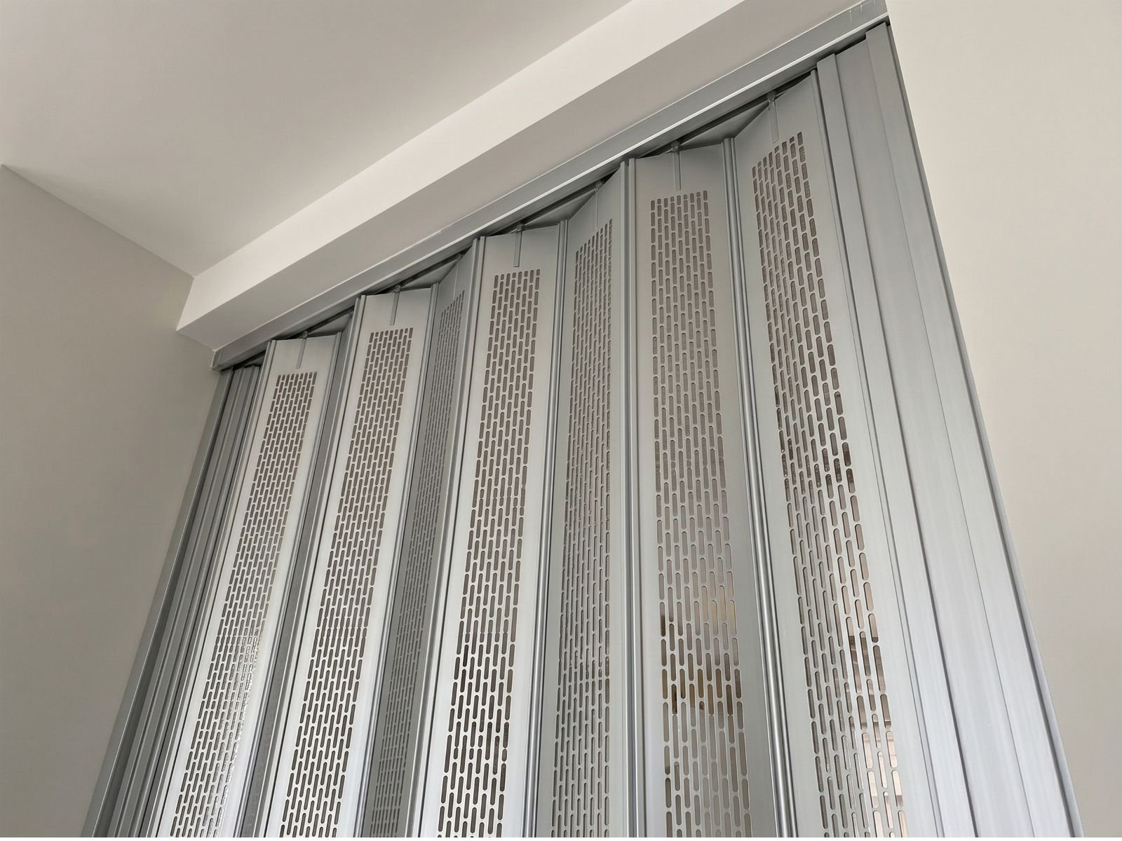 Un primer plano técnico que muestra un panel de la cortina de aluminio troquelada Modelo Aíra, con su patrón de listones oblongos de 6mm por 32mm, integrado en una estructura modular ligera de perfiles de aluminio y fijaciones de acero galvanizado.
